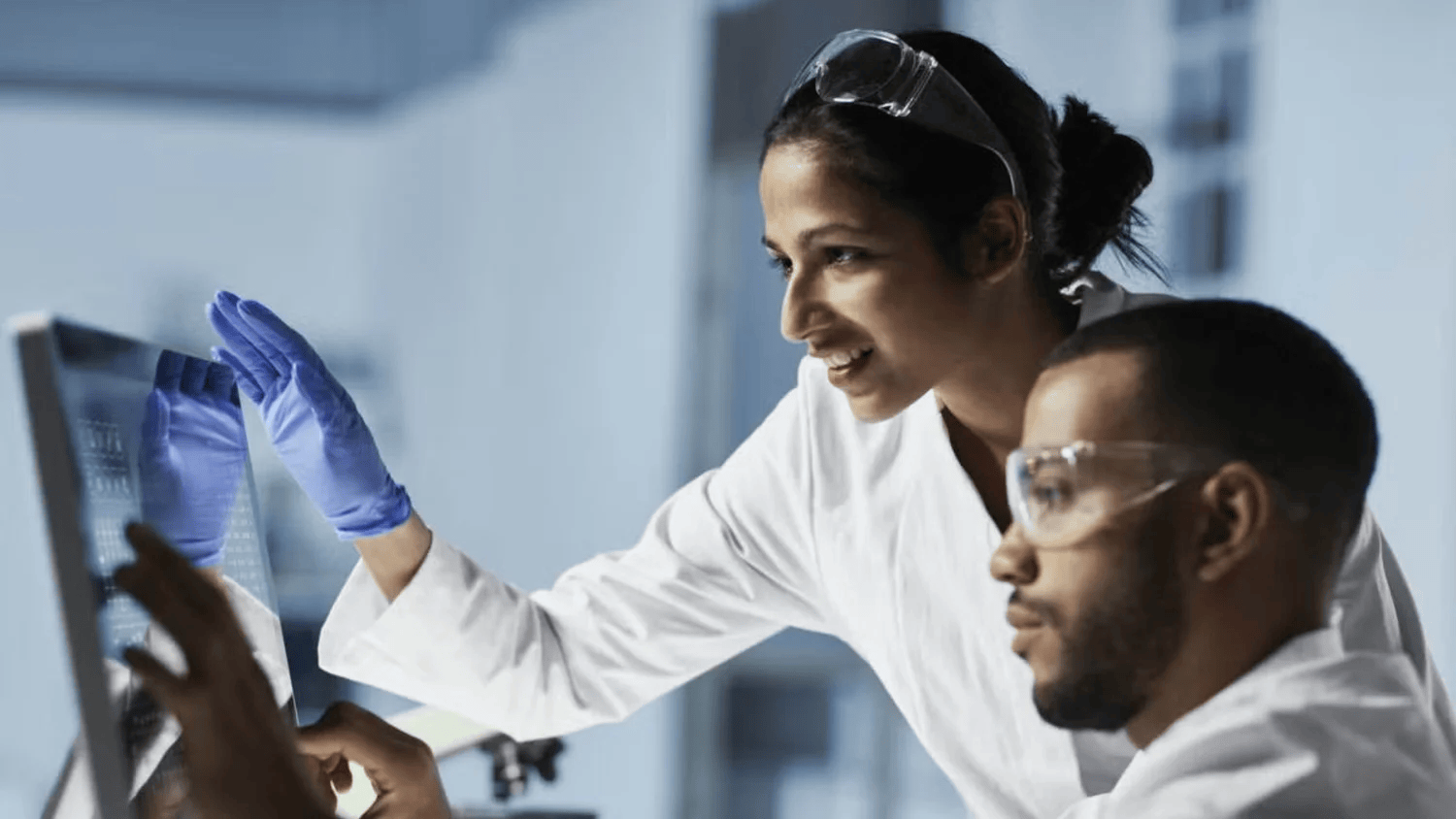 Female medical scientist smiling and looking at research on a monitor with male scientist wearing goggles