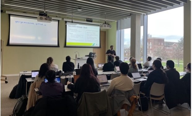 Group of diverse age and ethnicity students and researchers in a lecture room facing the boards with a male presenter presenting to them